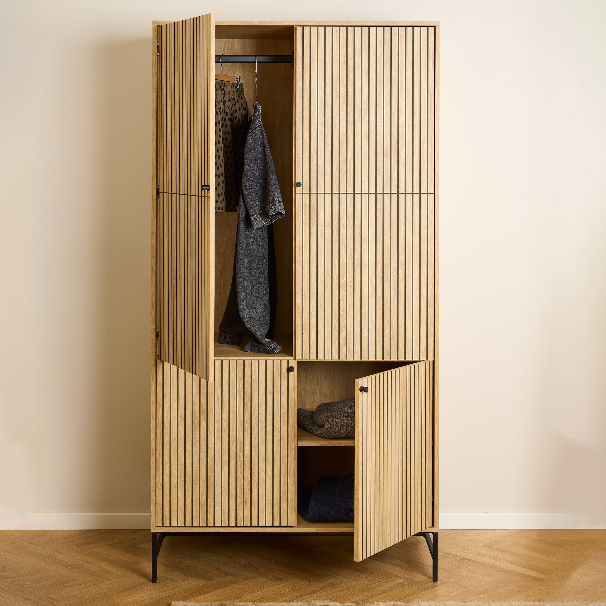 Large oak wardrobe with 4 vertically slatted doors and black steel base, opened to reveal hanging space and shelves, styled in a modern Scandinavian bedroom.