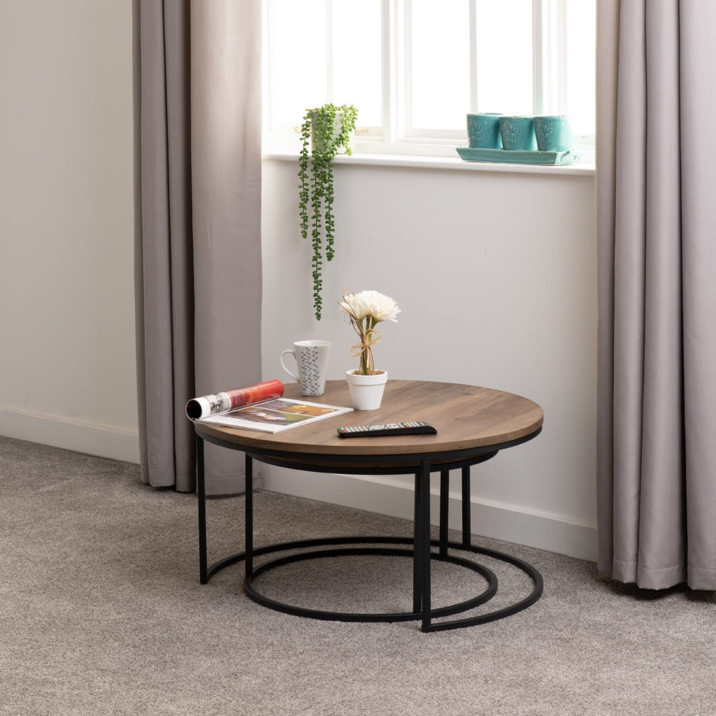 Quebec Round Coffee Table Set featuring two nesting tables with medium oak-effect tops and black metal frames, styled in a bright living room with neutral curtains and minimalist decor.