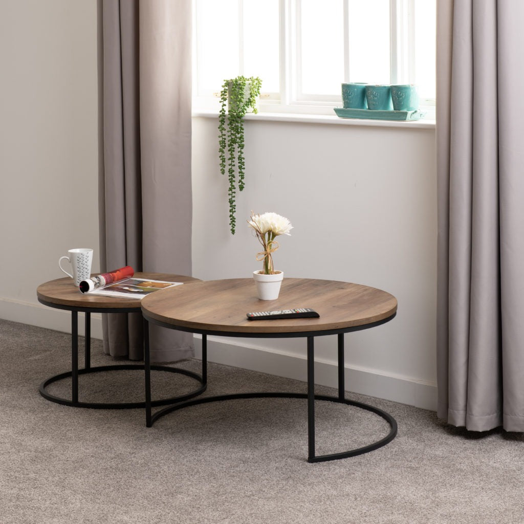 Quebec Round Coffee Table Set featuring two nesting tables with medium oak-effect tops and black metal frames, styled in a bright living room with neutral curtains and minimalist decor.
